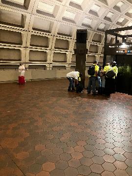 A picture of the elevator from the upper level to the lower level. Beyond the elevator, which is to the right, there is a wall. There are construction workers in front of the elevator working on the structure.